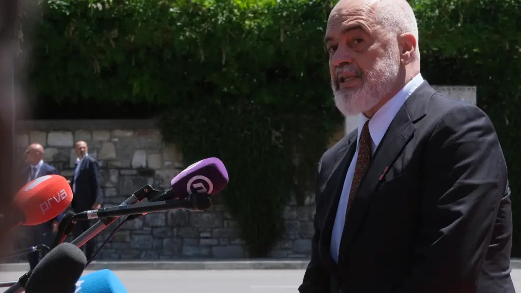 Bald man with a gray beard in a dark suit and patterned tie speaks to the press outdoors, microphones in front.