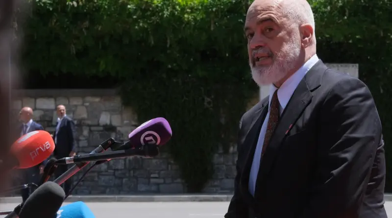 Bald man with a gray beard in a dark suit and patterned tie speaks to the press outdoors, microphones in front.