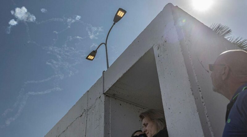 People stand at a white concrete shelter under a blue sky with a streetlight above and sun glare to the right, a man wearing sunglasses looking toward the opening.