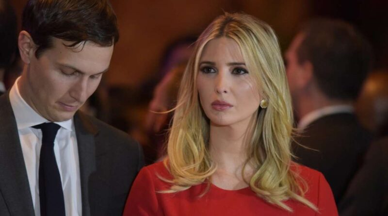 Man in a dark suit and tie looks down beside a blonde woman in a red dress at a formal event.