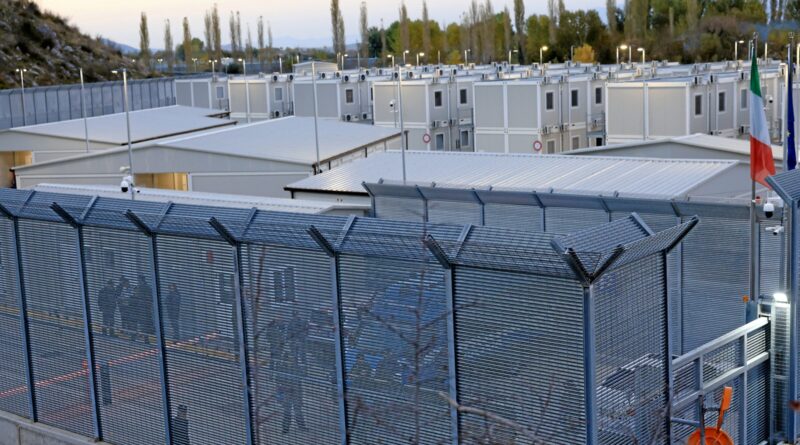 Row of prefabricated white modular buildings in a secure industrial complex, with metal fencing in the foreground and Italian and EU flags nearby.