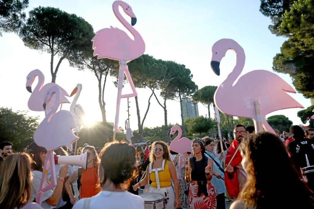 Crowd at an outdoor festival holding pink flamingo cutouts on sticks, with trees and sun flare in the background.
