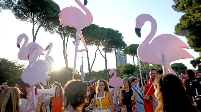 Crowd at an outdoor festival holding pink flamingo cutouts on sticks, with trees and sun flare in the background.