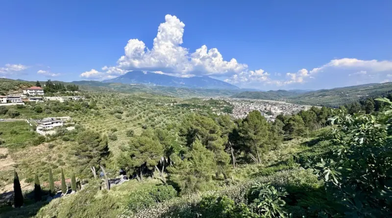 Wide landscape: hillside with trees and scattered houses, a town in the valley, and distant mountains under a blue sky with fluffy clouds.