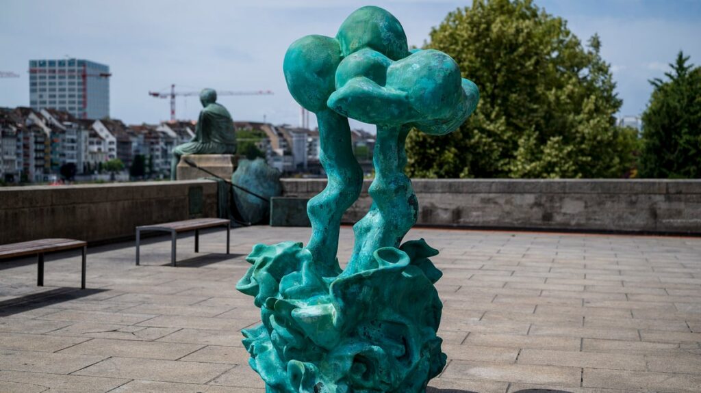 Turquoise abstract sculpture with twisting trunks rising from an ornate base in an open plaza, with benches and city buildings in the background.