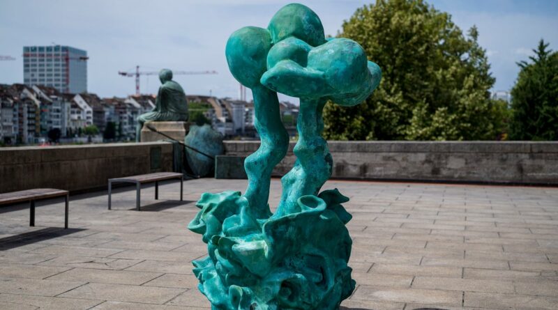 Turquoise abstract sculpture with twisting trunks rising from an ornate base in an open plaza, with benches and city buildings in the background.