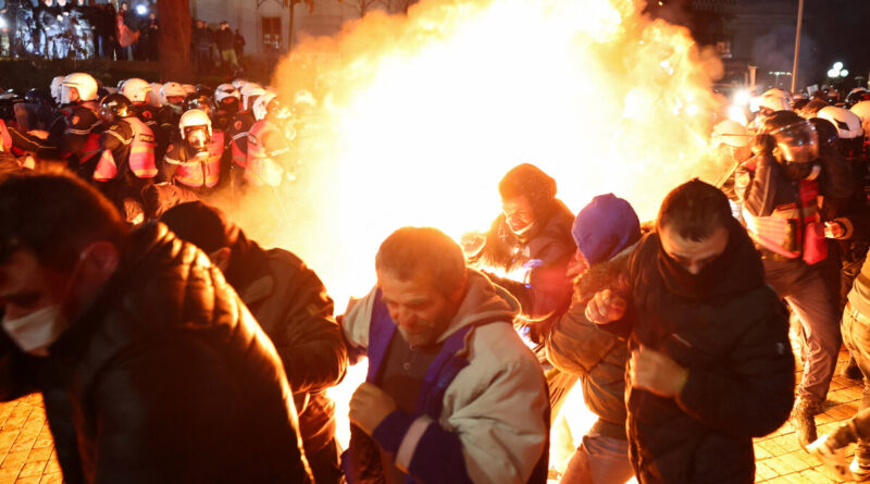 Crowd of protesters gathered at night around a large bonfire, flames lighting the scene as people move away from the fire.