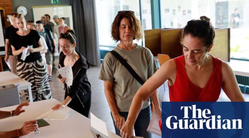 People line up to vote at a polling station, placing ballots on a table inside a bright room with large windows in the background.