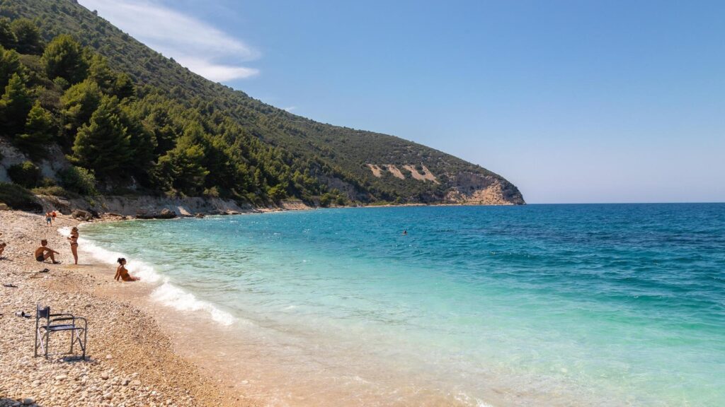 Pebble beach with turquoise sea, forested hillside on left, a few people relaxing near the water's edge and a metal chair on the shore