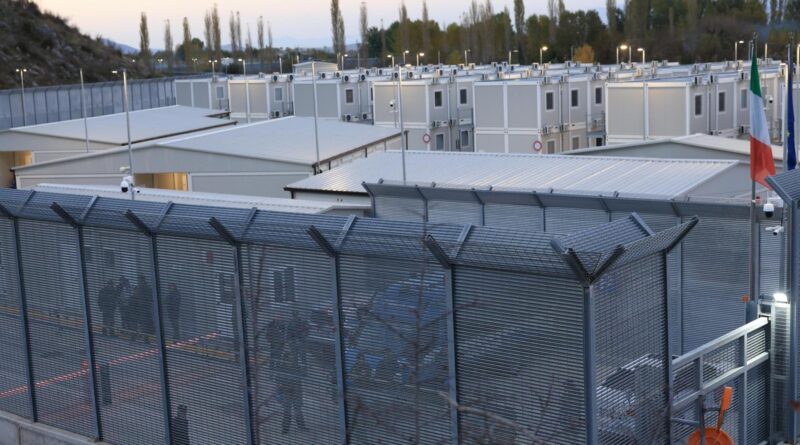 Secure compound with grey modular buildings behind tall angled metal fencing and visible flags (Italy and EU).