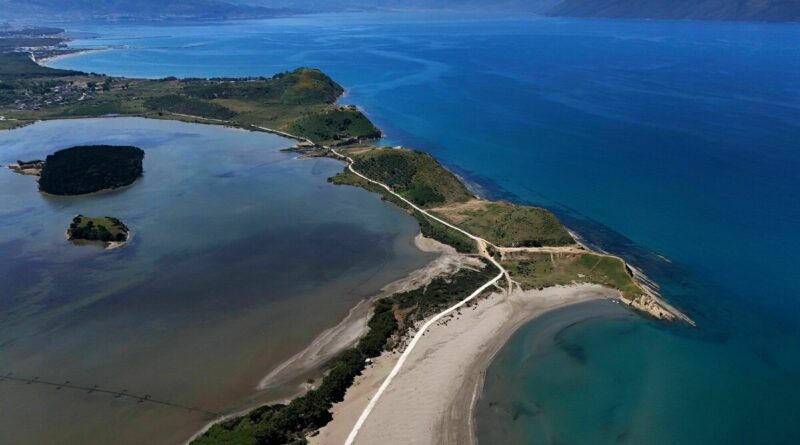 Aerial view of a narrow sandy peninsula connected by a white road to mainland, flanked by calm lagoons on the left and turquoise sea on the right, with distant mountains on the horizon.