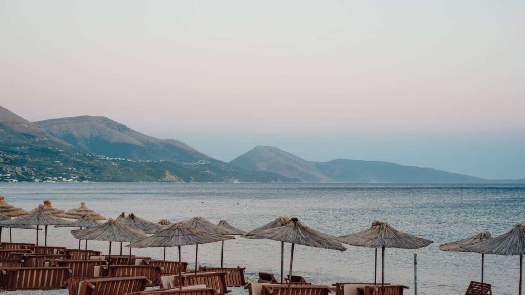 Row of straw beach umbrellas and wooden lounge chairs along calm sea with mountains in the distance at sunset.