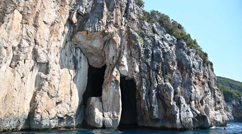 Coastal rock cliff with a dark sea cave at the water's edge under a clear blue sky.