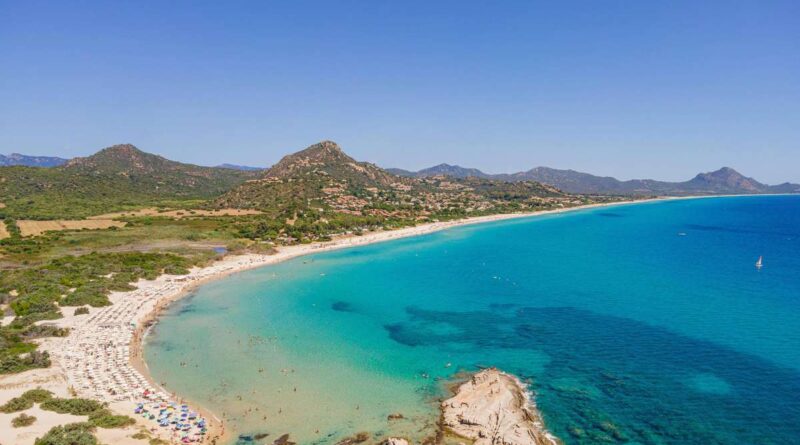 A curved sandy beach with turquoise water, sun umbrellas, and green hills in the distance under a clear blue sky.