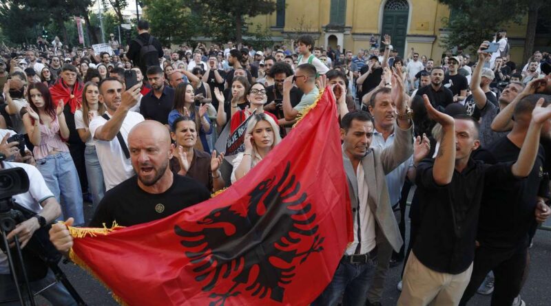 Crowd of protesters in a street, several people raise hands while one man holds a red Albanian flag with a black eagle in the foreground.