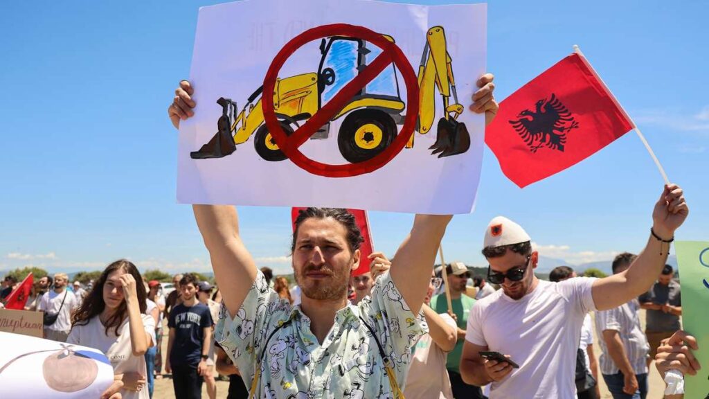 Protesters march outdoors; man holds a sign with a drawing of a construction excavator crossed out, while another person waves an Albanian flag nearby.