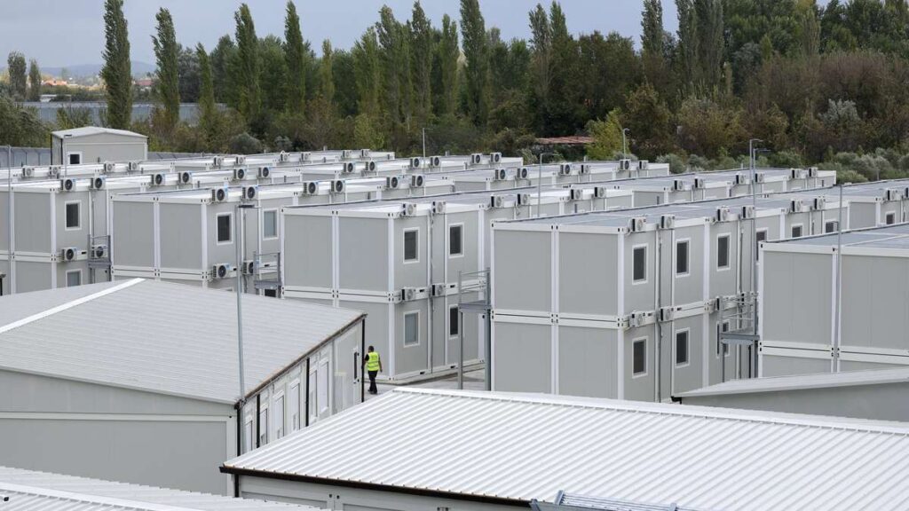 Rows of gray modular container buildings forming a temporary housing site, with a worker in a yellow safety vest walking between units; trees in the background.