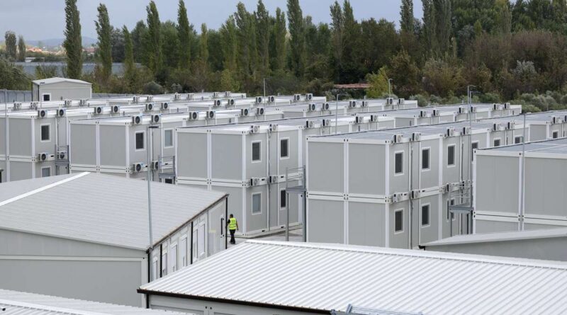 Rows of gray modular container buildings forming a temporary housing site, with a worker in a yellow safety vest walking between units; trees in the background.
