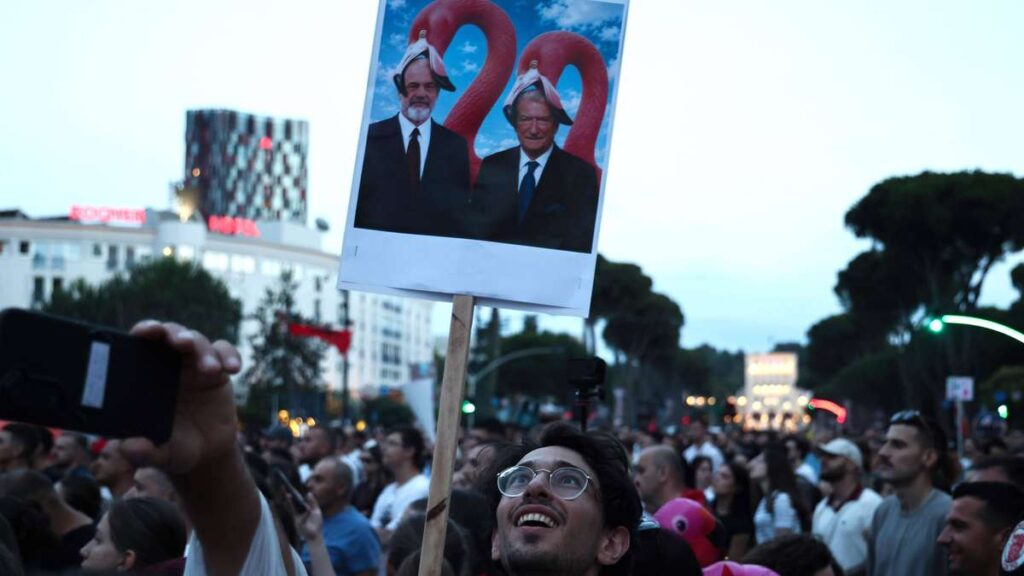 Crowd at dusk protest; a person holds up a sign showing two suited men with flamingo headgear against a blue sky image.