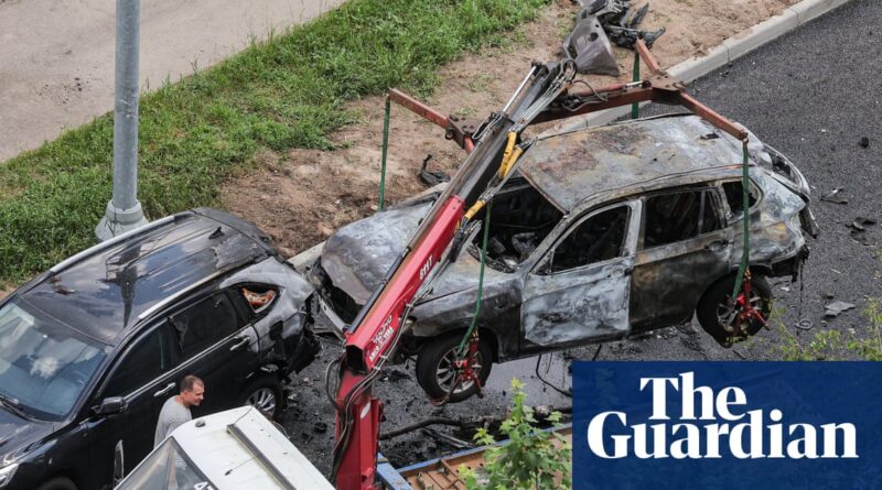 Two severely burned cars on a roadway being lifted by a tow crane for removal, with a Guardian logo overlay in the corner.
