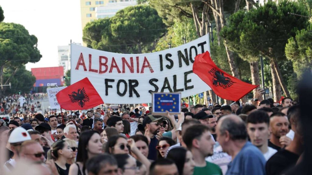 Protest crowd holding a large banner reading 'ALBANIA IS NOT FOR SALE' with red Albanian flags and a blue EU flag in front of trees and buildings, daytime urban rally.