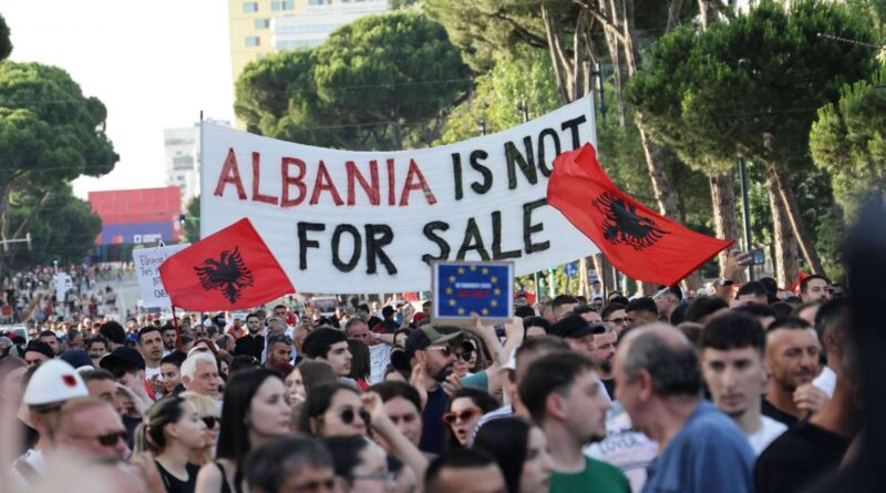 Protest crowd holding a large banner reading 'ALBANIA IS NOT FOR SALE' with red Albanian flags and a blue EU flag in front of trees and buildings, daytime urban rally.