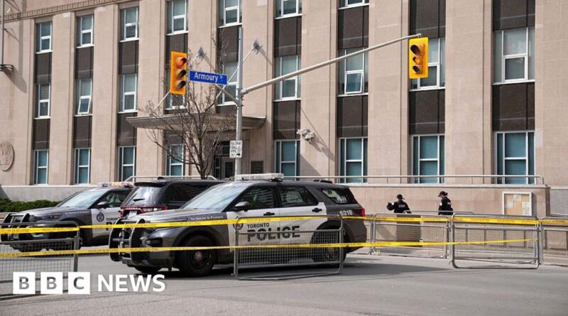 Police cars and yellow caution tape cordon off a city street in front of a large building, with officers in the background; BBC News watermark present.