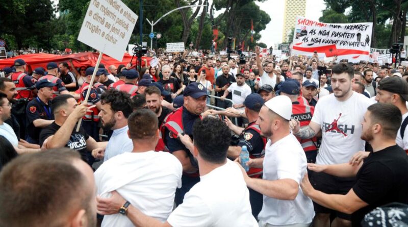 Large crowd of protesters pushing against police in red vests at a city street demonstration; signs visible in the background.