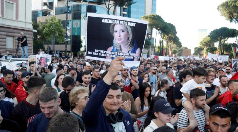 Large crowd of protesters in an urban street, a man in the foreground holds a placard with a portraits and bold text headline.