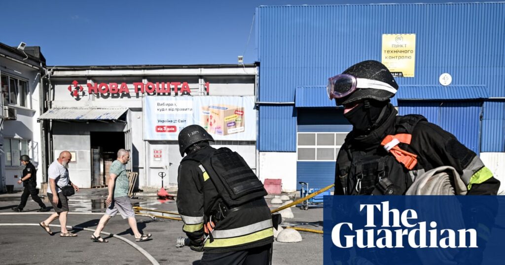 Firefighters in protective gear working outside a blue warehouse with a Nova Poshta sign on a sunny day, as pedestrians pass by.