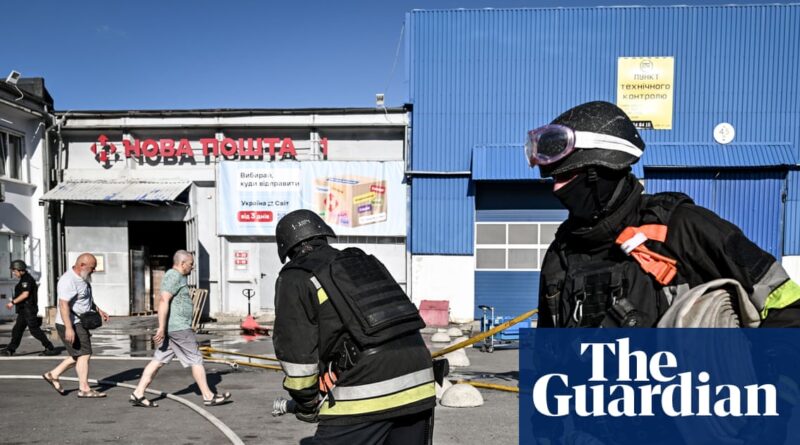 Firefighters in protective gear working outside a blue warehouse with a Nova Poshta sign on a sunny day, as pedestrians pass by.