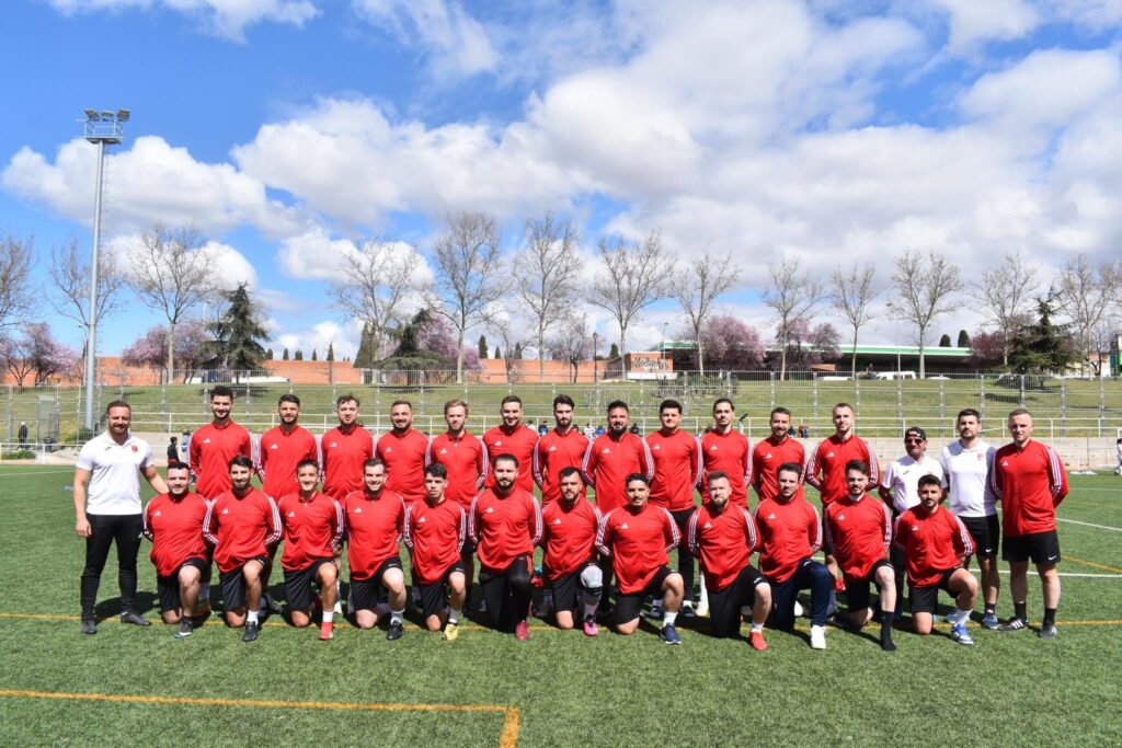 Soccer team in red jerseys posing for a group photo on a green field with a fence and blue sky behind them