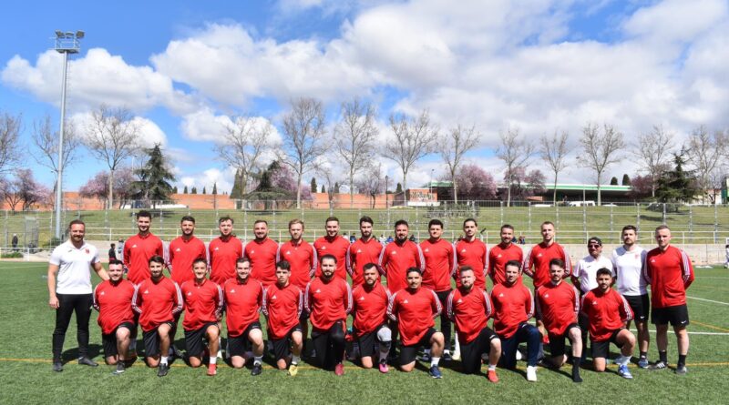 Soccer team in red jerseys posing for a group photo on a green field with a fence and blue sky behind them
