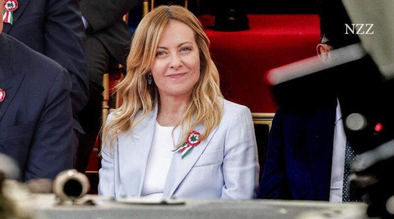 Woman in a light blue blazer at a formal event, smiling toward the camera; wearing a red‑green‑white ribbon pin on her lapel.