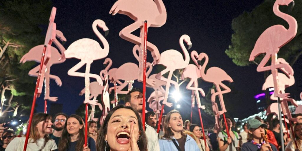 Crowd at night festival holding pink flamingo decorations on red poles, cheering.
