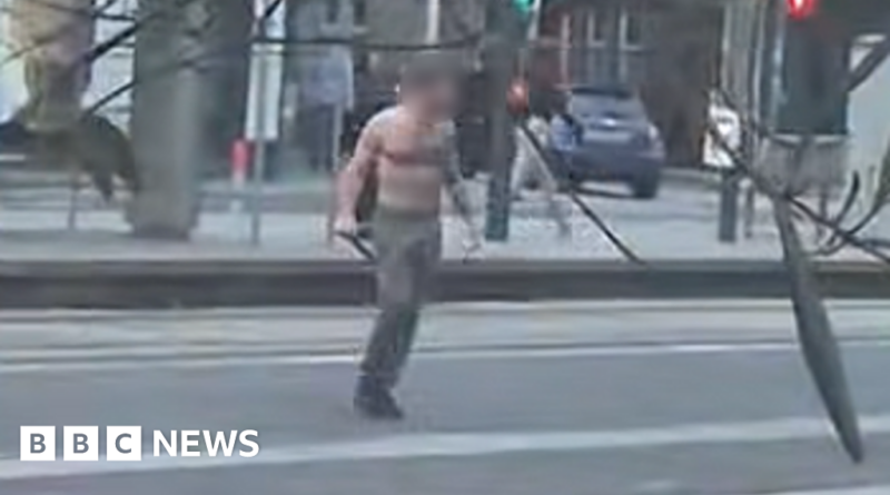 Shirtless man standing on a city street holding two long black poles, with a car and pedestrians in the blurred background (BBC News).