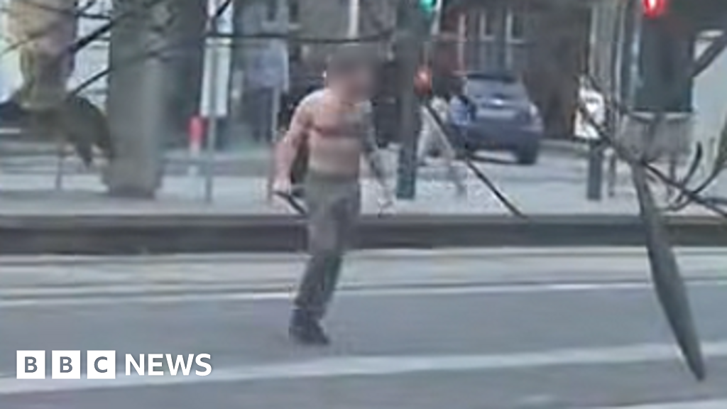 Shirtless man standing on a city street holding two long black poles, with a car and pedestrians in the blurred background (BBC News).