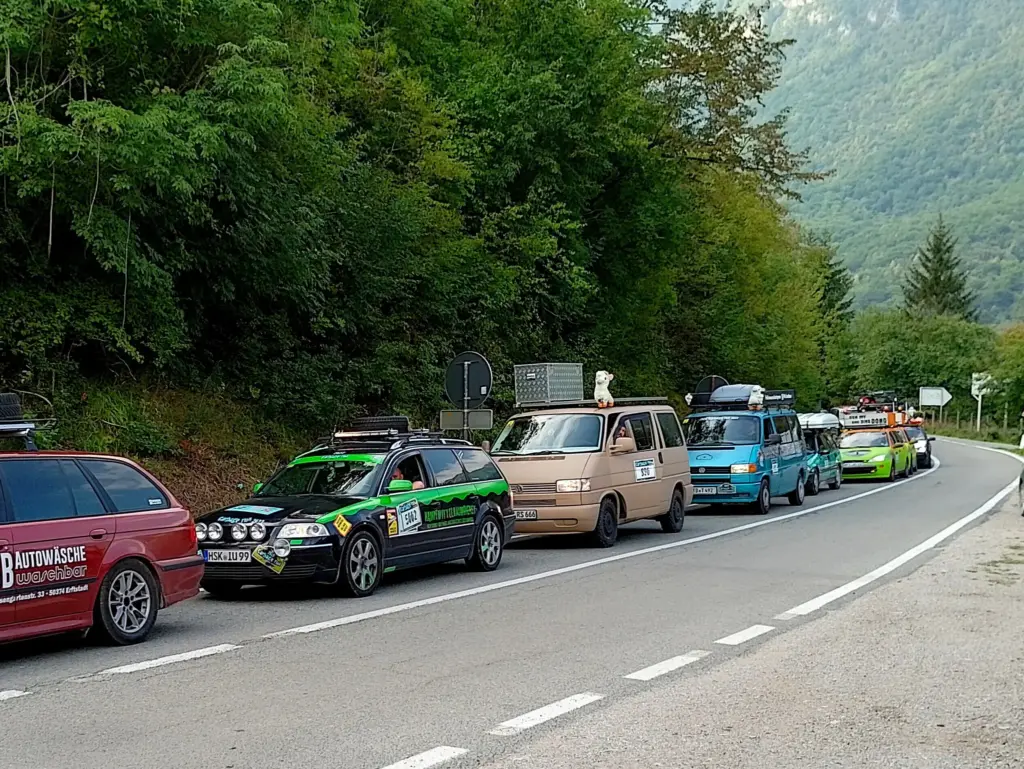 Line of decorated cars in a mountain road convoy beside dense green trees and distant hills/valley scenery.