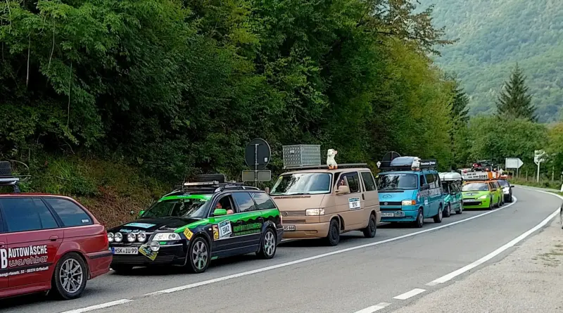 Line of decorated cars in a mountain road convoy beside dense green trees and distant hills/valley scenery.
