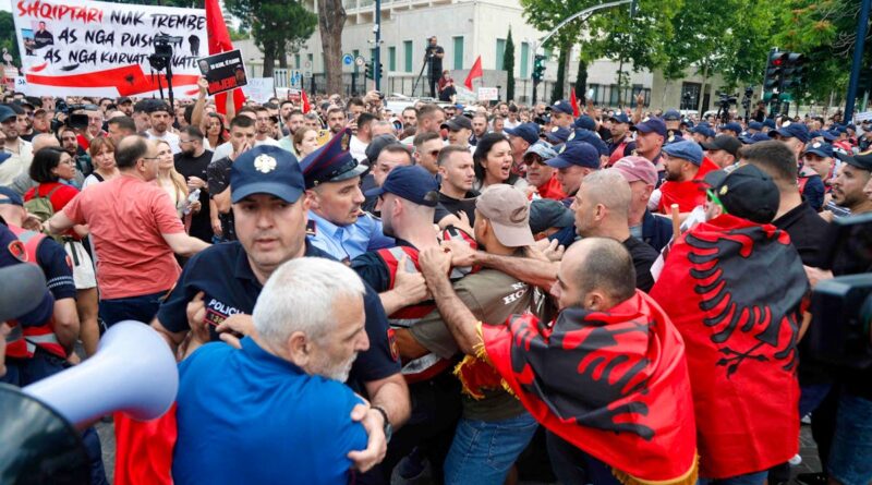 Crowded street protest with police restraining a man; banner and red flags visible in the background.
