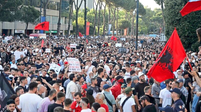 Large crowd of protesters filling a street, many holding signs with a red Albanian flag prominently visible above the crowd.