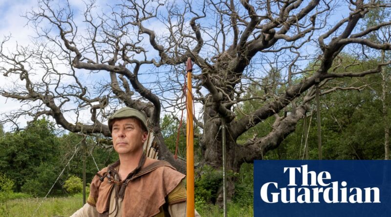 Man in medieval archer attire with a bow and arrows stands before a large bare-branched tree outdoors; Guardian logo overlayed on the bottom right.