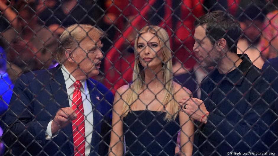 Three people stand behind a chain-link fence at a crowded event; a man in a suit with a red tie speaks to a blonde woman in a black dress, while another man looks on.