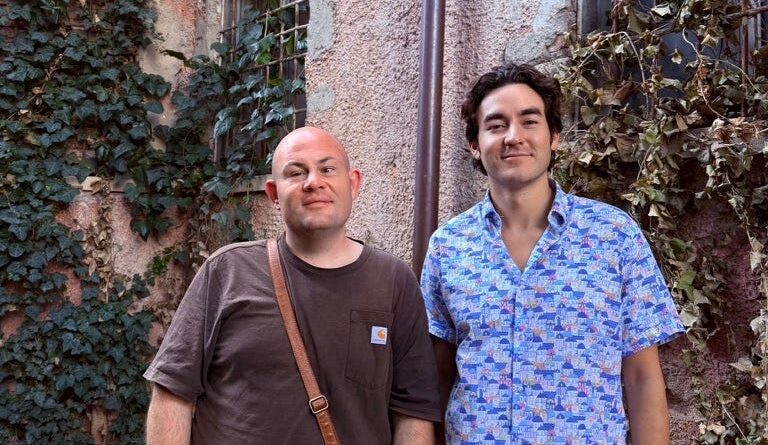 Two men stand side by side outdoors in front of ivy-covered walls, one bald wearing a brown shirt and crossbody bag, the other in a blue patterned shirt.