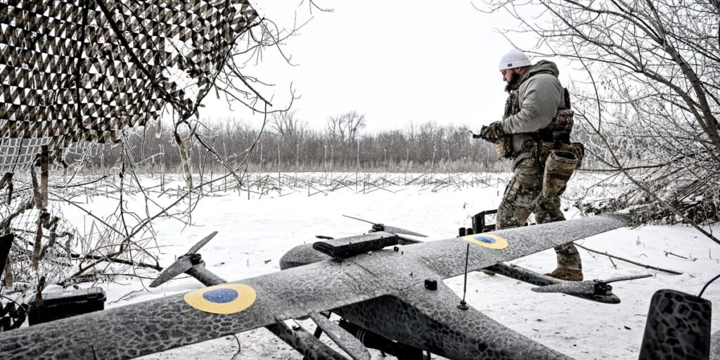 Soldier in winter camouflage stands next to a camouflaged unmanned aircraft on a snow-covered field.