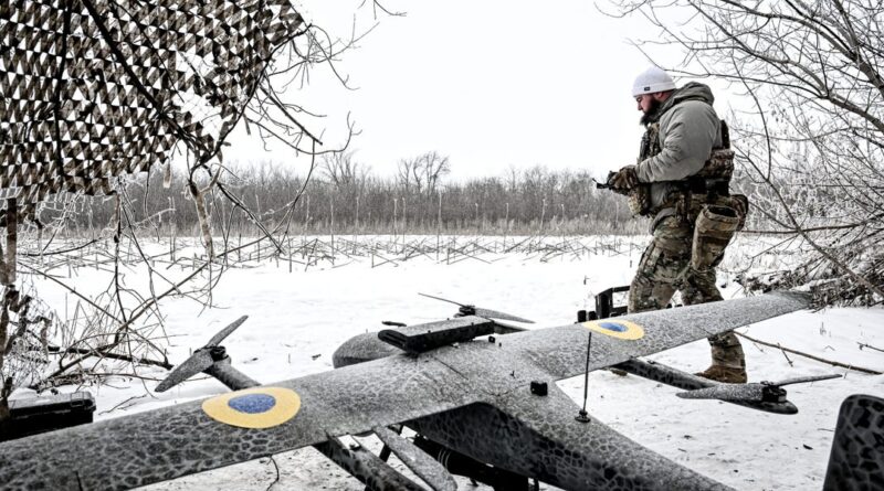 Soldier in winter camouflage stands next to a camouflaged unmanned aircraft on a snow-covered field.