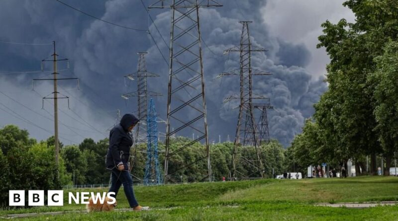 Person wearing a dark hoodie walks along a grassy area near tall power line towers as thick smoke rises in the distance, suggesting a fire nearby.