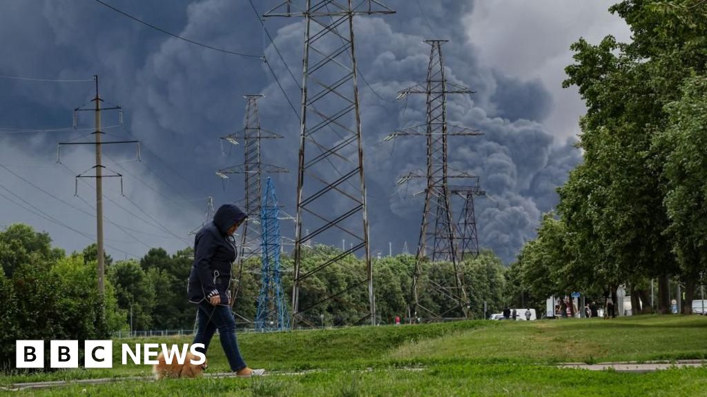 Person wearing a dark hoodie walks along a grassy area near tall power line towers as thick smoke rises in the distance, suggesting a fire nearby.