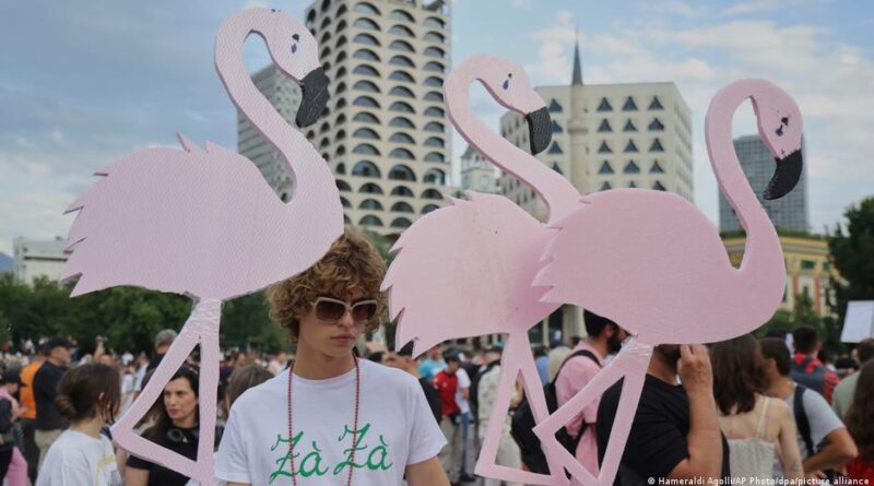 Two people hold oversized pink flamingo cutouts at a crowded outdoor festival in an urban square with modern buildings in the background.