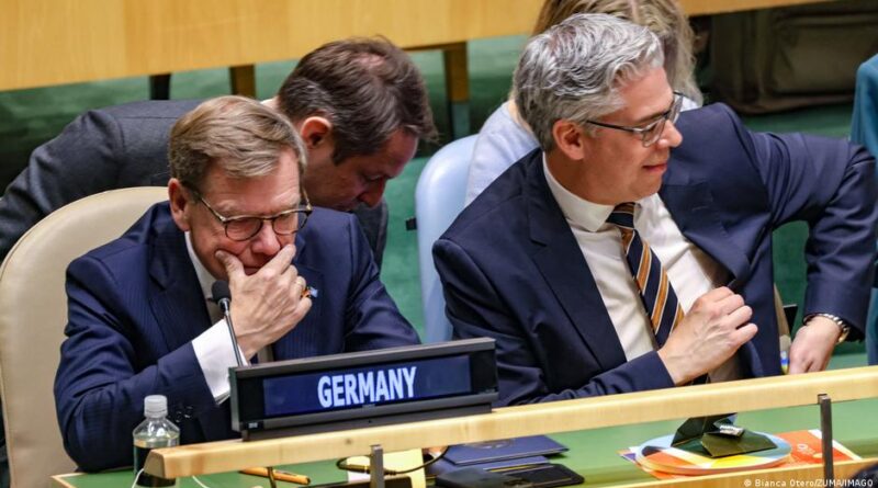 Two suited delegates sit at a conference table with a 'Germany' nameplate, one resting his chin on his hand as others look on, microphones and papers nearby.